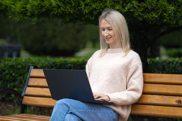 Side view of a young beautiful smiling blonde sitting on a bench with a laptop in the park.