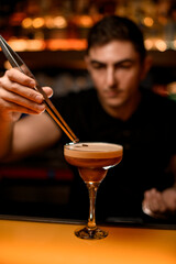glass with frothy espresso martini cocktail which bartender decorates with coffee bean using tweezers