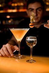 Barman holds glass with cold cocktail and garnishes it with physalis using tweezers