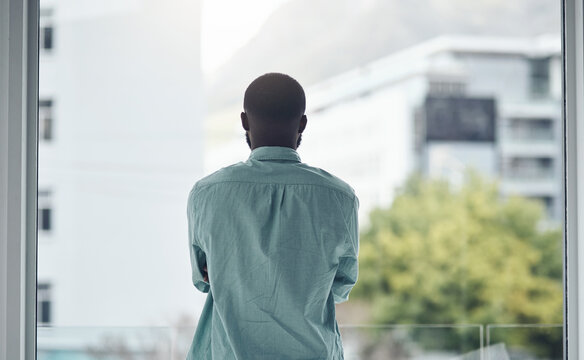 Back, thinking and window with a business black man standing in his office looking at a city view. Idea, motivation and inspiration with a male employee at work, contemplating a decision in town