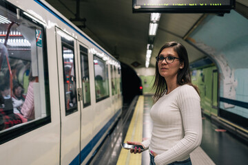 Young brunette woman waiting on the platform for the subway to stop while holding her mobile phone in her hand.