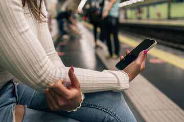 Unrecognizable young woman waiting on the platform sitting looking at her cell phone while waiting for the subway to arrive.