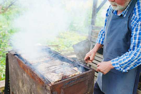 an elderly man with a beard in work clothes cooks meat steaks on the grill 