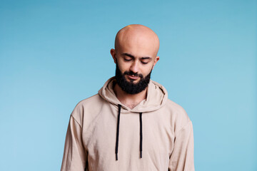 Arab man with neutral expression looking down and tilting head. Pensive young person wearing casual beige hoodie clothes thinking and standing in casual pose on blue background