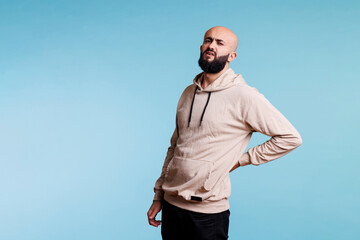 Fototapeta premium Arab man with lower back injury and looking at camera with painful facial expression. Grimacing young person suffering from backache and holding hand on sore muscles portrait