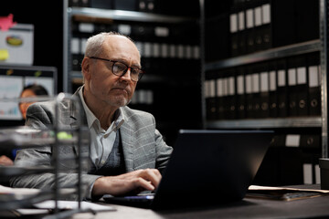 Senior private detective checking confidential criminology evidence files on laptop. Investigator in agency file storage office surrounded by folders on cabinet shelves - obrazy, fototapety, plakaty