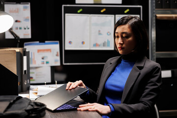 Asian businesswoman shutting down laptop at work preparing to go home. Bookkeeping consultant executive surrounded by accountancy file cabinet office statistics data flowcharts