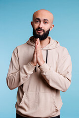 Arab man praying with folded palms and looking at camera, making wish with hopeful facial expression. Young adult person pleading and asking for forgiveness studio portrait