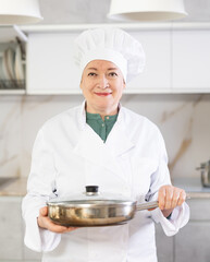 Smiling mature female chef standing in the home kitchen while cooking and holding a frying pan in her hands