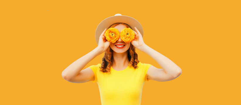 Summer portrait of happy smiling young woman covering her eyes with flower buds as binoculars looking for something wearing round straw hat on yellow background