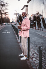 A stylish black bald man, with a long beard with grey hair, casually dressed in pink, holds a cup of coffee as he is about to cross the bicycle lane in the afternoon, he checks if the road is clear