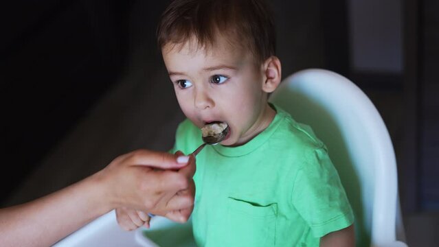 Toddler sitting at feeding table eats from fork. Mom’s hand gives a spoon with porridge to her child. Top view close up.