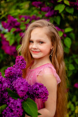Fototapeta premium a beautiful happy smiling blonde girl with long hair in a pink dress with a purple bouquet of lilacs in her hands stands against the background of a lilac bush