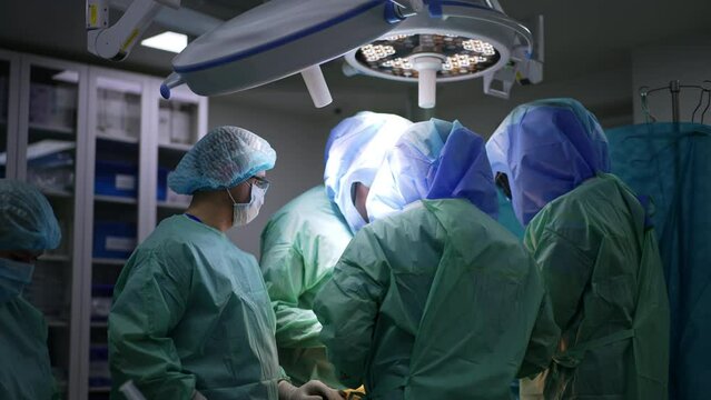 Group of surgeons in protective suits and helmets work under the round lamps in surgery room. Team of doctors cooperate at work.