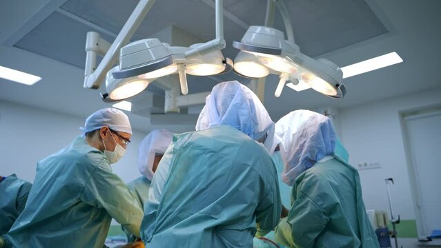 Large surgical team is busy at operational room. Doctors in protective suits stand around the patient on surgery table.