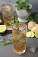 Glass of tasty ginger ale with ice cubes and ingredients on grey wooden table