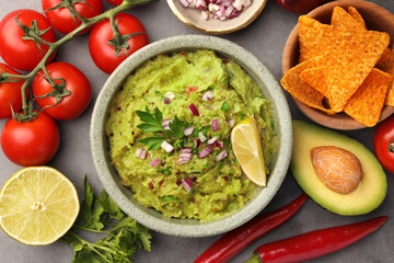 Bowl of delicious guacamole, nachos chips and ingredients on grey table, flat lay