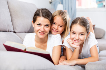 Positive woman reading book to two kids resting on couch in living room