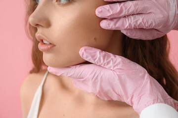 Dermatologist examining moles on young woman's face against pink background, closeup