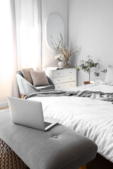 Interior of light bedroom with modern laptop on bedside bench and blooming tree branches