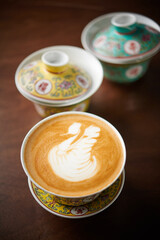 Hot latte art cappuccino in a unique and antique chinese cup with some cups as background.