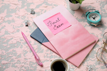 Sticky note with text FINAL EXAM, notebooks, alarm clock and cup of coffee on grunge pink background