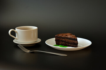 A piece of chocolate puff cake with mint leaves on a black background, and a cup of hot coffee on a saucer with a fork.