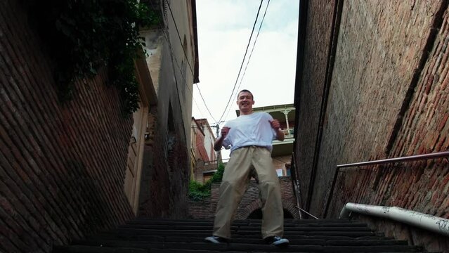 A guy in a white T-shirt defiantly dances a modern dance on the streets of the old city. Historical streets of Tbilisi. A young dancer improvises in a dance and steps up the steps. Freestyle, hip hop.