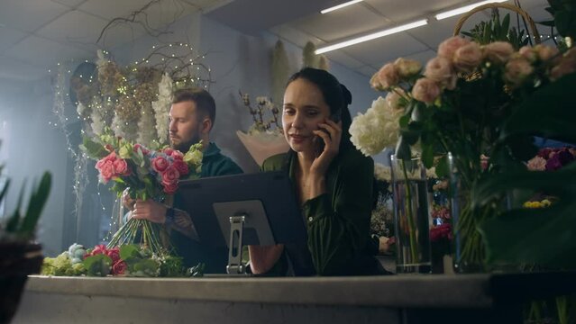 Female entrepreneur, florist talks by phone with customer and takes online order in flower store using digital tablet. Male coworker collects bouquet. Concept of small business and entrepreneurship.