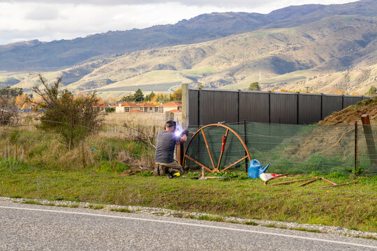 Man diligently welds metal construction in an outdoor setting. Surrounded by fields, mountains, and a sky adorned with clouds,