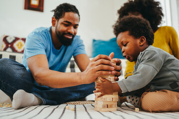 An african american family is spending time at home and playing a Jenga game together while sitting on the floor.