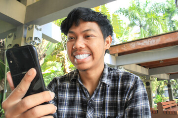 young asian man smilling with white tooth while looking at black mobile phone feeling happy and...