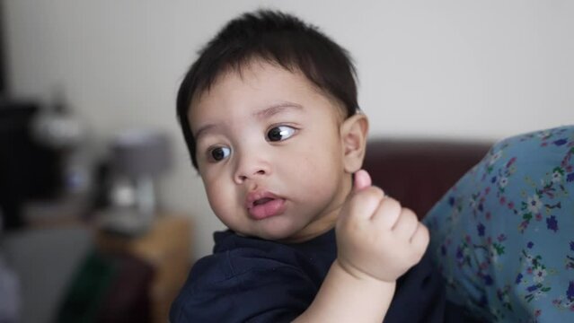 A happy young Asian baby boy being held in his mother’s arms as he chews on food. Slow Motion