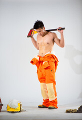 Topless professional firefighter with a sweaty body is standing on a white background holding an iron axe on his shoulder while looking at the floor.