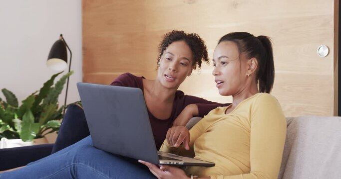 Happy biracial lesbian couple relaxing on couch together, using laptop and talking, in slow motion