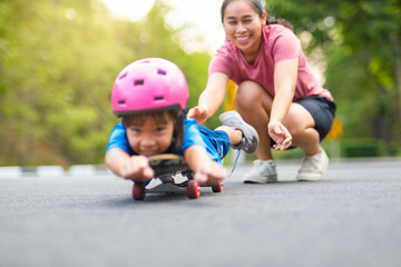 Fototapeta na wymiar Active little girl and mom enjoy skateboarding. Cute little girl wearing helmet practicing skateboarding in park. Mother trains her daughter to skateboard. Outdoor sports for children.