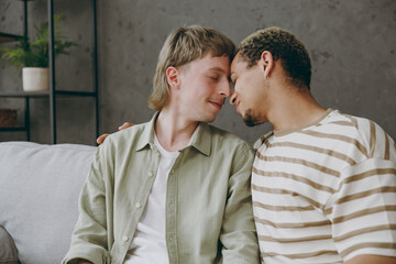 Young happy couple two gay men wear casual clothes together touch forehead close eyes sit on sofa couch at home flat rest spend free spare time in living room. Pride day june month love lgbtq concept.