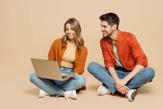 Full body young couple two friends family IT man woman wear casual clothes together sitting hold use work on laptop pc computer isolated on pastel plain light beige color background studio portrait.