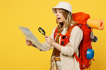 Surprised young woman carry backpack with stuff mat hold map use magnifier isolated on plain yellow...