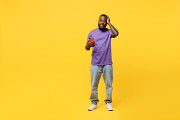 Fototapeta na wymiar Full body young man of African American ethnicity wear casual clothes purple t-shirt hold head use mobile cell phone lok camera isolated on plain yellow background studio portrait. Lifestyle concept.