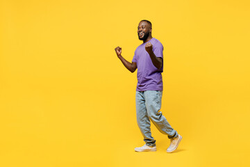 Fototapeta na wymiar Full body sideways young man of African American ethnicity wear casual clothes purple t-shirt walk go doing winner gesture celebrate clenching fists say yes isolated on plain yellow background studio.
