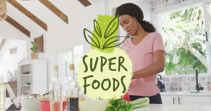 Animation of leaves and super foods text over african american woman cutting vegetables in kitchen