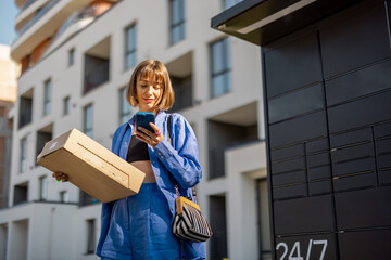 Young woman using smart phone while standing with a parcel delivered with post office machine with...