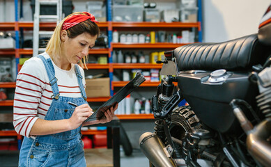 Concentrated mechanic woman with clipboard checking motorcycle and taking notes on factory