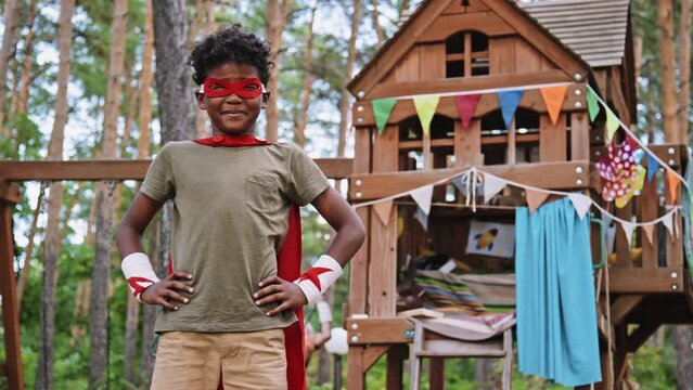 Portrait of African American elementary school boy posing in superhero costume in front of treehouse with playground