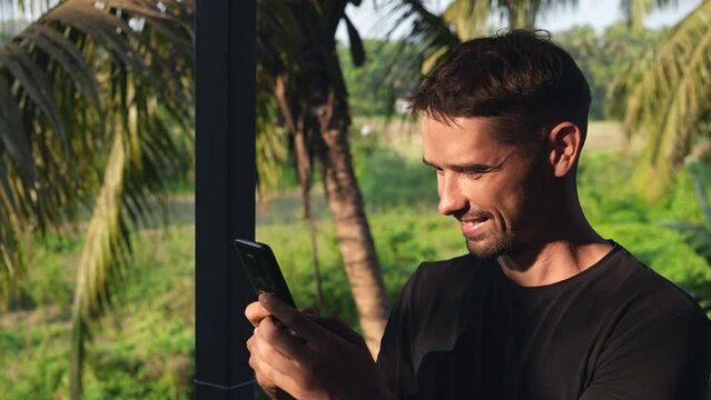 Happy male tourist write text message. Smiling man chatting. Tropical palm tree. Joyful guy typing sms close up. Person hold mobile cell phone. Social media. Sunny day relax trip. Use messenger device
