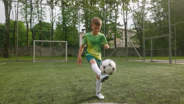 The boy dressed in a football uniform is practicing on the soccer field. The young footballer is training to juggle kick up the ball with his foot.