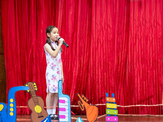 The Asian kid girl sing a song on stage at her school activity day, dress in Qipao style, red curtain background