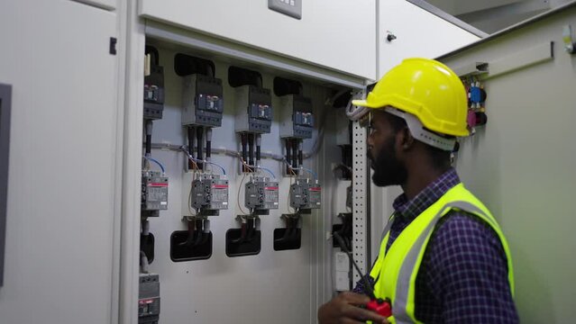 Professional African male engineer in safety uniform working at factory server electric control panel room. Industrial technician worker maintenance checking power system at manufacturing plant room.