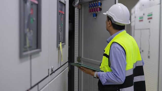 Professional Asian male engineer in safety uniform working at factory server electric control panel room. Industrial technician worker maintenance checking power system at manufacturing plant room.